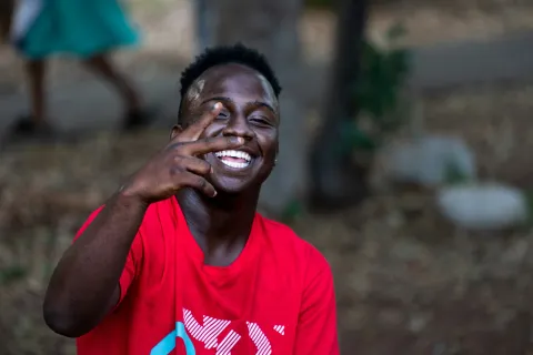 Smiling young man in a red shirt making a peace sign outdoors with blurred background.