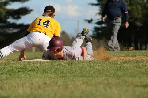 Baseball player in maroon slides headfirst into base while player in yellow attempts a tag during game.