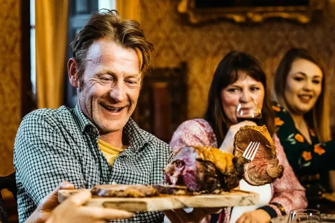 Group of people enjoying a meal with carved roast beef and wine at a dinner table in a warm setting