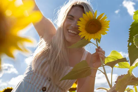 Smiling woman in striped dress holding sunflower in a bright field under a blue sky with clouds.