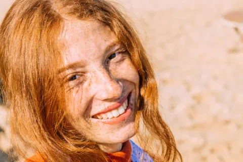 Close-up of a smiling red-haired woman with freckles on a sunny sandy beach background
