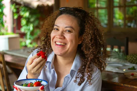 Smiling woman with curly hair eating fresh strawberries at a wooden table in a bright indoor setting.