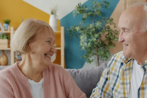 Smiling elderly couple sitting on a couch in a colorful living room, sharing a happy moment together.