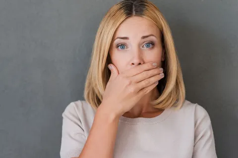 A woman with blonde hair covers her mouth, showing a surprised expression against a gray background.