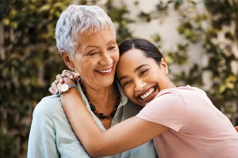 Happy young woman hugging older woman outdoors, both smiling warmly with greenery background.