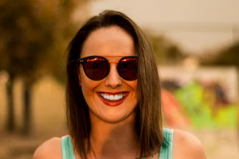 Smiling woman with sunglasses, wearing a light blue tank top, outdoors with a blurred colorful background.