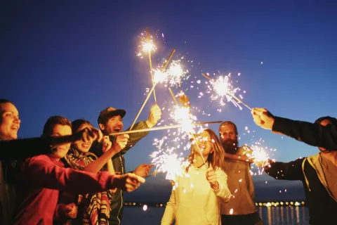Group of friends joyfully holding sparklers together at dusk near water with city lights in background