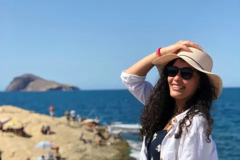 Woman in sunglasses and sunhat smiling by the rocky sea shore on a sunny day with ocean and island in background