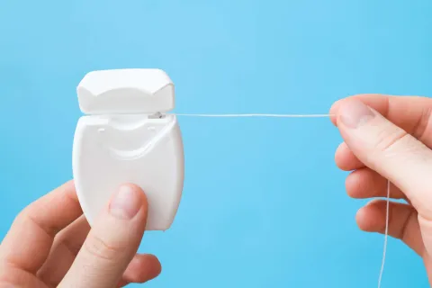 Close-up of hands holding dental floss container with floss being pulled out against a blue background.
