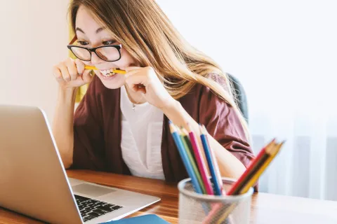 Frustrated young woman biting a pencil while working on a laptop at a bright desk with colored pencils.