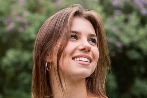 Smiling young woman with brown hair and hoop earrings outdoors with greenery and purple flowers behind her