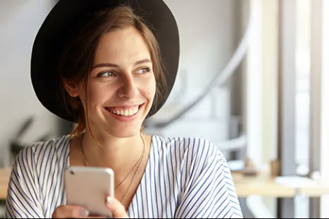 Young woman wearing a hat smiling and holding a smartphone while looking away in a bright indoor setting