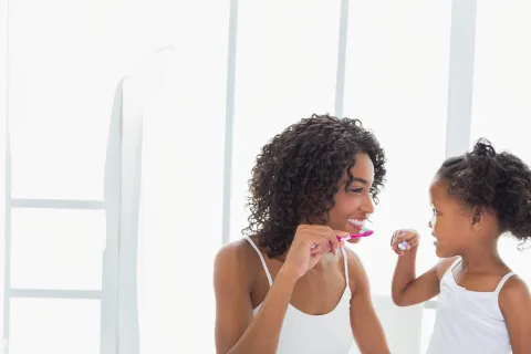 Mother and daughter smiling and brushing their teeth together in a bright bathroom setting.