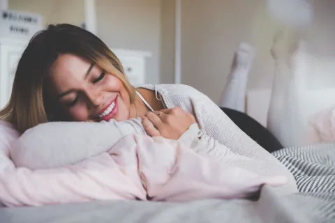 Smiling young woman lying on bed resting on her arm with cozy pillows and casual sweater indoors