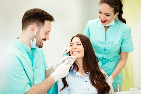 Dentist and assistant smiling while examining a female patient's teeth with a shade guide in a dental clinic.