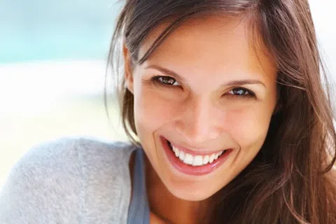 Close-up of a smiling young woman with long brown hair and bright eyes in natural light.