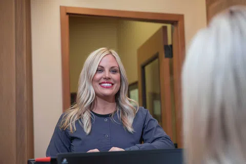 Smiling blonde woman with long hair wearing navy blue scrubs stands behind reception desk in office.