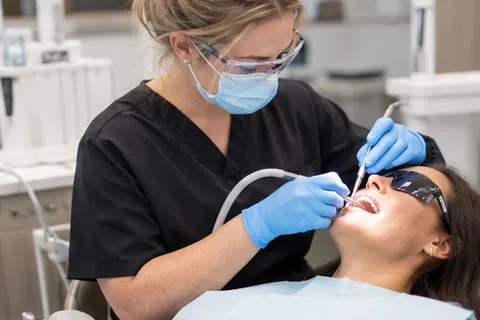 Dentist wearing mask and gloves examining a female patient's teeth in a bright dental clinic.