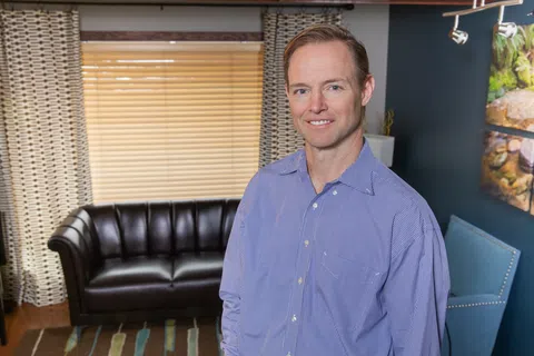 Smiling man wearing a black jacket with embroidered name Dr. Clayton and a white shirt indoors.