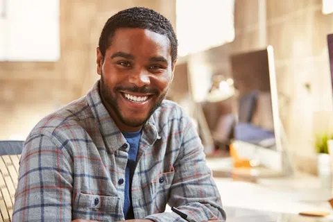 Smiling man in plaid shirt sitting at a desk in a bright modern office with computer and plants