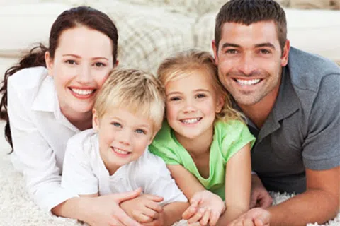 Happy family of four with parents and children smiling and lying on a carpet indoors.
