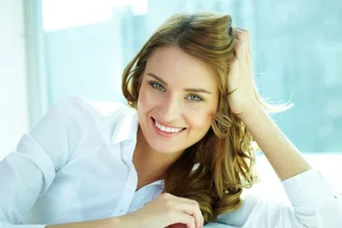 Smiling woman with long hair wearing a white shirt reclining comfortably by a window with natural light.