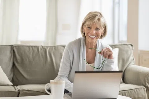 Smiling mature woman holding glasses using laptop on couch in bright living room with coffee mug nearby