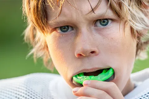 Close-up of a young athlete putting a green mouthguard in his mouth, wearing sports gear outdoors.