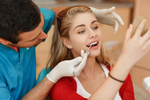Dentist examining a smiling young woman’s teeth while she looks into a handheld mirror during a dental checkup.