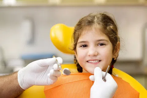 Smiling young girl in an orange bib sitting in a dental chair with dentist's gloved hands holding tools.