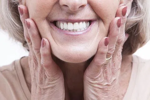 Close-up of an elderly woman smiling with hands gently on her cheeks showing white teeth and natural wrinkles