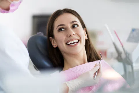 Smiling woman at dental clinic sitting in chair while dentist in gloves holds dental tool.