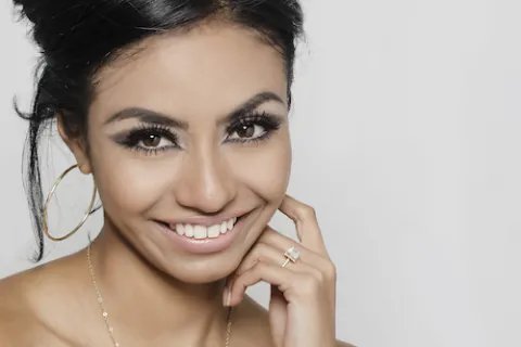 Smiling woman with dark hair, large hoop earrings, and a sparkling ring against a plain light background.