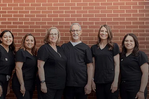 Smiling medical team in black scrubs standing against a brick wall background in a group portrait.
