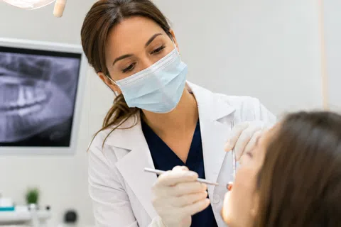 Dentist wearing a mask examining a patient's teeth using dental tools with dental X-ray in background