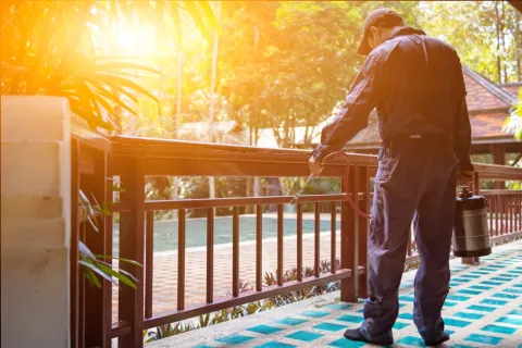 Man in protective clothing spraying insecticide on outdoor wooden railing during daytime.