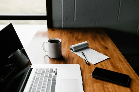 Workspace with laptop, white coffee mug, notepad with pen, and smartphone on wooden table by window.