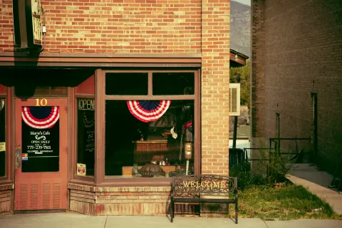 Exterior of a brick rural small business cafe with patriotic red, white, and blue bunting, a welcome bench, and an open sign.