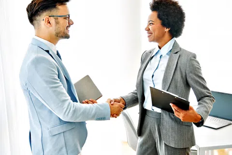 Two business professionals shaking hands in office, smiling and holding tablet and clipboard, wearing suits.