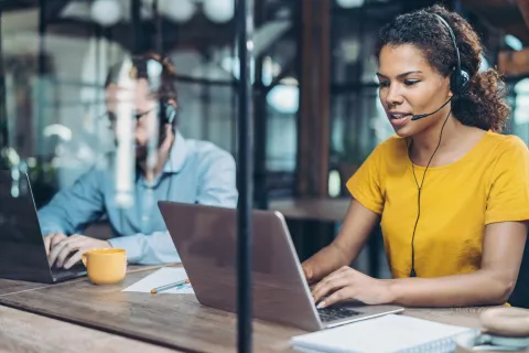 Woman in yellow shirt wearing headset working on laptop in office with male colleague in background.