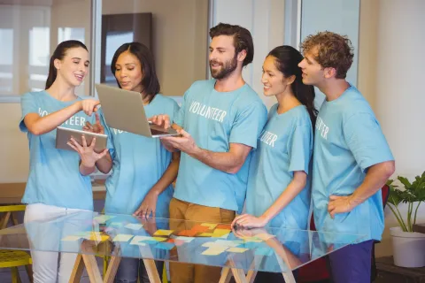 Five volunteers in blue shirts collaborate using laptop and tablet at a table covered with sticky notes.