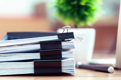 Stack of thick documents secured with black binder clips on a wooden desk near a laptop and pen.
