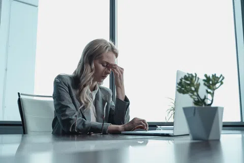 Woman in business attire sitting at desk with laptop, looking stressed and rubbing her forehead in office.