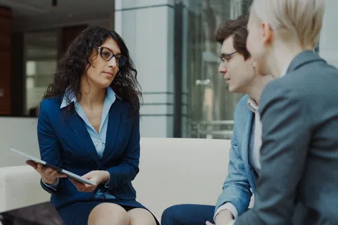 Businesswoman in navy suit holding documents consulting two colleagues in an office lobby sitting on a white sofa.
