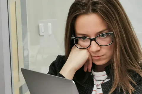Focused woman wearing glasses working on a laptop in an office setting with notebook and coffee mug nearby