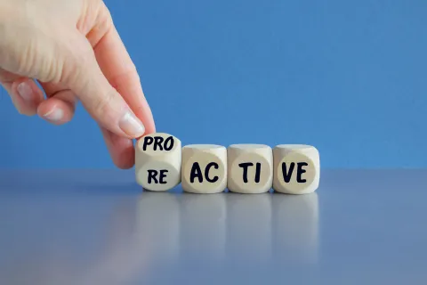 Hand flipping a wooden block from RE to PRO in the word REACTIVE and PROACTIVE against blue background.