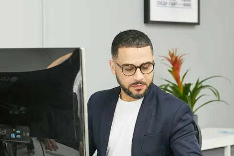 Man in glasses and blazer writing notes at desk with computer and plant in modern office.