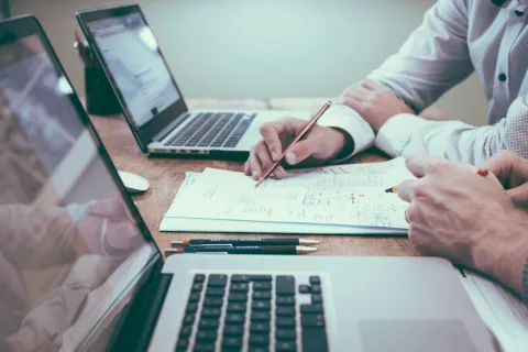 Two professionals collaborate over documents and laptops, planning and analyzing data at a wooden desk.