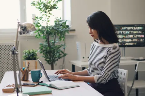 Woman in striped shirt working on laptop at a desk with a plant and notebook in a bright office.