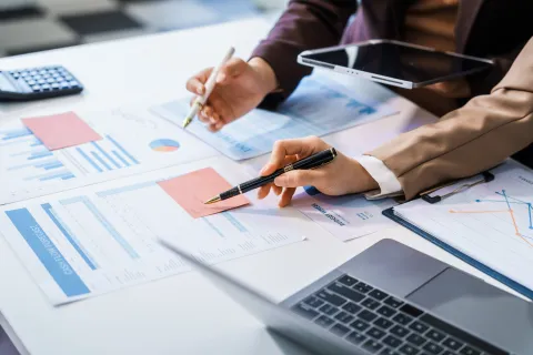 Two professionals analyzing charts and graphs with pens, tablet, and laptop on white desk in office.
